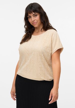 Kobieta o falowanych ciemnych włosach, ubrana w beżowy koronkowy top z krótkim rękawem i czarną plisowaną spódnicę, stojąca na białym tle.