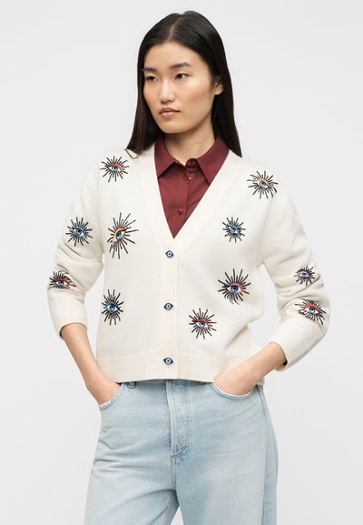 Λευκό πλεκτό ζακέτα με κεντημένα μοτίβα ήλιου και ματιού, μπλε κουμπιά και ριμπ μανσέτες. Συνδυασμένο με μακρυμάνικο πουκάμισο μπουρντό και ανοιχτόχρωμα τζιν.