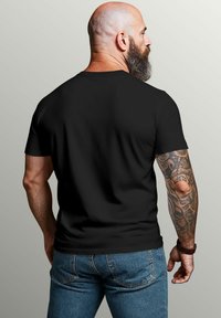 Schwarzes T-Shirt mit kurzen Ärmeln und Rundhalsausschnitt, von hinten an einem Mann präsentiert. Er hat einen vollbärtigen Bart und Tattoos auf seinen Armen. Denim-Jeans sind sichtbar.