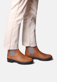 Botas Chelsea de cuero marrón con paneles laterales elásticos que presentan rayas azul y blanca, suela de goma negra y acabado texturizado.