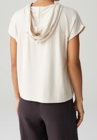 Jasno beżowa bluzka z drapowaną kapturem, krótkimi rękawami i prostym dołem, w połączeniu z ciemnymi spodniami. Gładka tekstura z minimalną liczbą szwów.