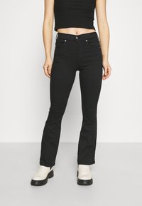 Jeans pretos de corte fluido feitos de denim, com cintura média, cinco bolsos e acessórios metálicos. Combinados com botas brancas até o tornozelo.