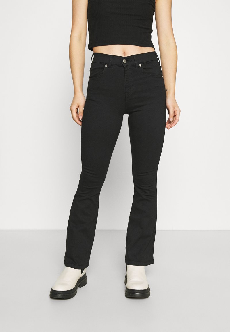 Jeans pretos de corte fluido feitos de denim, com cintura média, cinco bolsos e acessórios metálicos. Combinados com botas brancas até o tornozelo.