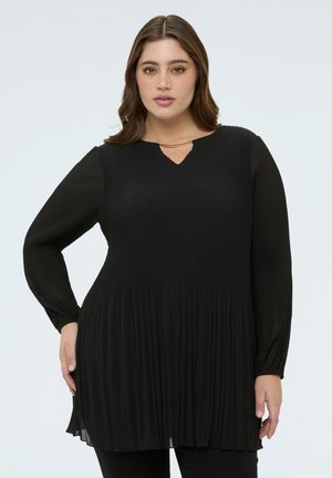 Kvinde med langt brunt hår iført en sort plisseret tunika med lange ærmer, en lille V-udskæring og et guldhalskæde mod en ensfarvet baggrund.