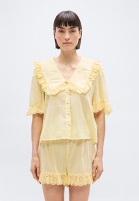 Femme portant un chemisier boutonné jaune pâle avec col et manches à volants, assorti à un short à volants, debout devant un fond blanc.