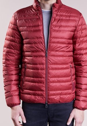 Mężczyzna ubrany w czerwoną pikowaną kurtkę puchową na jasnoszarym swetrze, w czarnych spodniach, stoi na jednolitym tle.