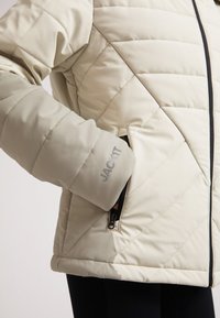 Beige kviltet jakke med høj krave, der har skrå syninger, en lynlåslomme og et logo på ærmet. Glat tekstur.