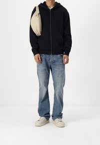 Sudadera con capucha negra con cremallera, vaqueros azul claro, bolso bandolera beige, zapatillas crema. Atuendo casual con un ajuste relajado y diseño minimalista.