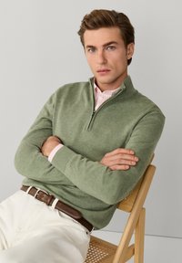 Młody mężczyzna siedzący na drewnianym krześle z plecionym siedziskiem, ubrany w zielony sweter na zamek na różowej koszuli w paski oraz białe spodnie z brązowym paskiem.