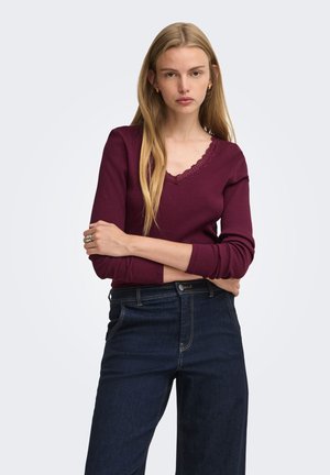 Młoda kobieta o długich blond włosach, ubrana w bordową bluzkę z długim rękawem i koronkowym wykończeniem oraz ciemnoniebieskie jeansy z wysokim stanem, stojąca z założonymi rękami.