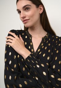 Blusa nera con motivo a pois dorati, tessuto leggero, scollatura profonda e polsini elasticizzati con dettaglio arricciato.