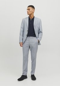 Lichtblauw op maat gemaakt pak met een marineblauwe poloshirt. Het pak heeft een enkelvoudige sluiting, revers met inkepingen en zijzakken. Zwarte schoenen.