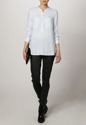 Femme portant une tunique bleu clair boutonnée, pantalon slim noir, bottines noires, tenant une pochette noire, debout sur un fond uni.