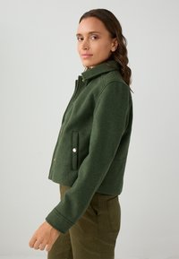 Giacca in misto lana verde con colletto alto, bottoni a pressione e due tasche laterali, abbinata a pantaloni verde oliva. Texture liscia e vestibilità su misura.