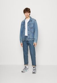 Svetle modrá džínsová bunda s knoflíkovým zapínáním vpředu a dvěma náprsními kapsami, kombinovaná s tmavými džíny a vícebarevnými teniskami. Pod ní je obyčejné bílé tričko.