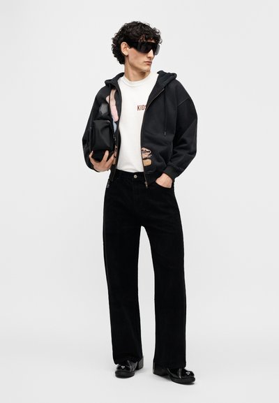 Μαύρο φούτερ με φερμουάρ και γραφικά, λευκό μπλουζάκι με κείμενο "KIDS", μαύρο παντελόνι κοτλέ, γυαλιστερά παπούτσια και μεγάλα μαύρα γυαλιά ηλίου.