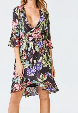 Vrouw draagt een zwarte bloemen wikkeljurk met een diepe V-hals en 3/4 mouwtjes met ruches, gestrikt in de taille met een bijpassende riem.