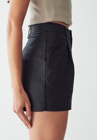 Černé lněné šortky s vysokým pasem, precizním střihem, bočními kapsami a jemným skladem na přední straně. Hladká textura a čisté linie.