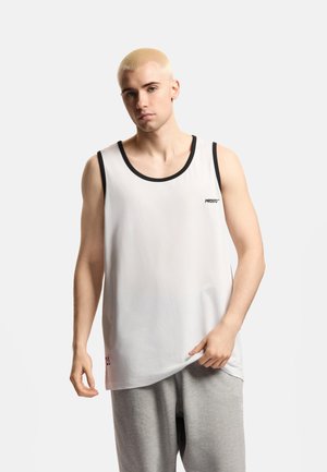 Młody mężczyzna z krótkimi, blond włosami, ubrany w biały sportowy bezrękawnik z czarnym wykończeniem i szare spodnie dresowe, stojący na jasnym tle.