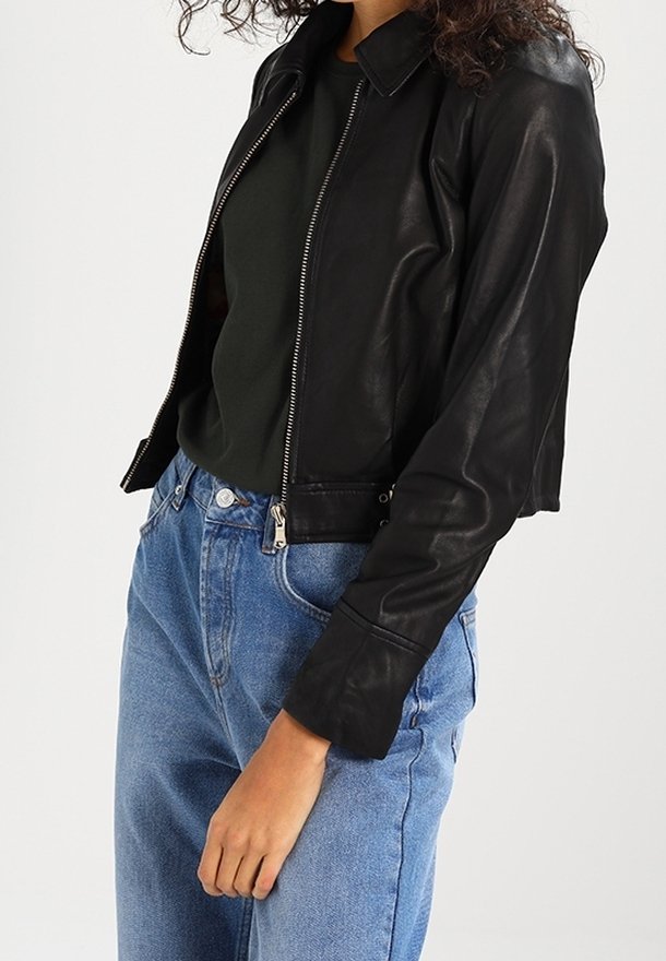 Femme portant une veste en cuir noire par-dessus un top noir avec un jean bleu taille haute, debout devant un fond uni.