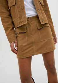 Veste en daim brun avec jupe assortie, dotée de poches avant et de coutures blanches contrastantes, mettant en valeur une coupe ajustée et une texture lisse.