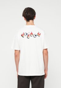 T-shirt bianca in cotone con una grafica colorata "VOLCOM" con fiamme e occhi sul retro. Maniche corte e vestibilità comoda.