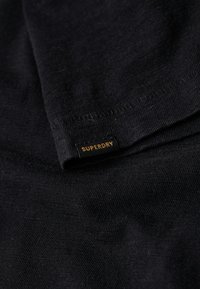 Czarna bawełniana koszulka z gładką fakturą, z małą metką "SUPERDRY" w czerni i złocie na dolnym brzegu.