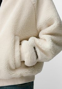 Chaqueta de forro polar color crema con material texturizado. Presenta un puño acanalado con un logo negro. Superficie suave y peluda, con un diseño de corte relajado.