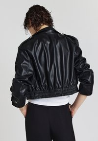 Schwarze, aus Kunstleder gefertigte, cropped Jacke mit tailliertem Schnitt, Schulterepauletten und gerafften Ärmeln. Glatte Oberfläche mit glänzendem Finish.