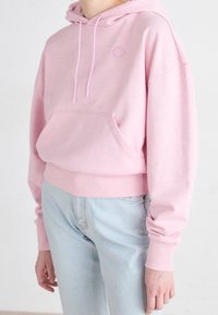 Sudadera rosa con un bolsillo tipo canguro, capucha con cordón, puños y bajo acanalados. Lleva unos vaqueros de mezclilla azul claros. Logo sutil en el pecho.