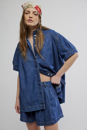 Pitkätukkainen nainen, jolla on kukkakuviollinen päähine, yllään ylisuurikokoinen sininen denim-paitapusero ja siihen sopivat farkkushortsit, poseeraa yhdellä kädellä taskussa.