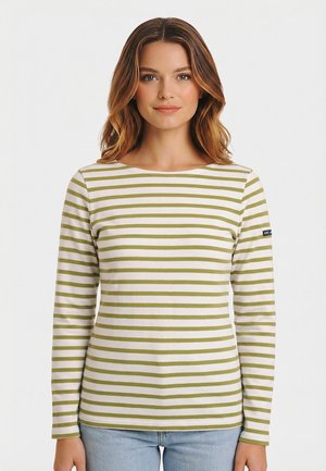 Donna con capelli castano ondulati che indossa una maglietta bianca a maniche lunghe con strisce orizzontali verde oliva e jeans azzurri, rivolta verso il fronte.