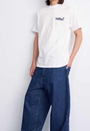 Personne portant un t-shirt blanc à manches courtes avec le texte "Wiltopia" et un jean bleu ample, debout avec une main dans la poche.