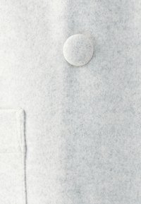 Tela de fieltro gris claro con una textura suave, que presenta un pequeño botón circular y un detalle de bolsillo cosido en el lado izquierdo.