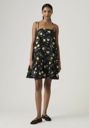 Frau steht und trägt ein schwarzes ärmelloses Kleid mit gelbem Blumenmuster sowie schwarze Ballerinas vor einem einfarbigen hellen Hintergrund.