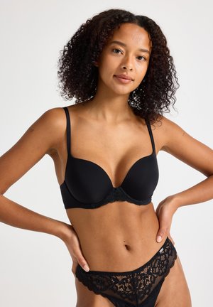 Ung kvinde med krøllet hår iført sort bh og blonder trusser, poserer med hænderne på hofterne mod ensfarvet baggrund.