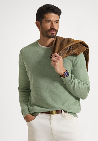 Light green knitted sweater with long sleeves, paired with cream-colored pants. Holding a brown leather jacket, wearing a blue watch.