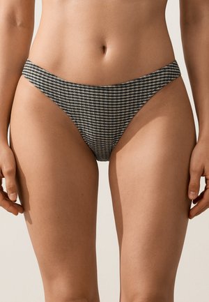 Femme portant un bas de bikini taille haute avec un motif à carreaux noir et blanc, debout devant un fond clair uni.