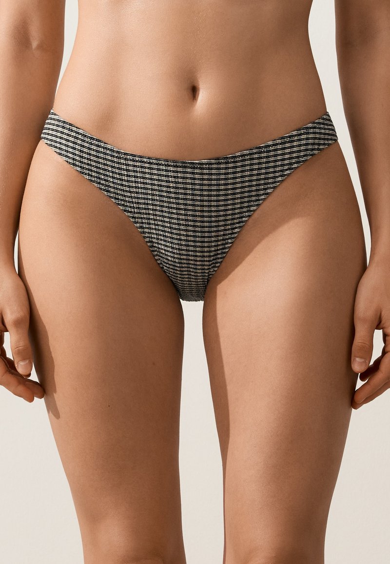 Femme portant un bas de bikini taille haute avec un motif à carreaux noir et blanc, debout devant un fond clair uni.