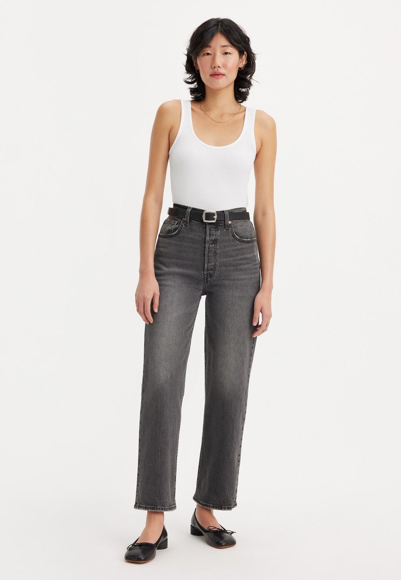 Jean noir à taille haute, coupe droite, avec une ceinture noire. Un débardeur blanc à côtes ajusté et des chaussures plates noires complètent le look.