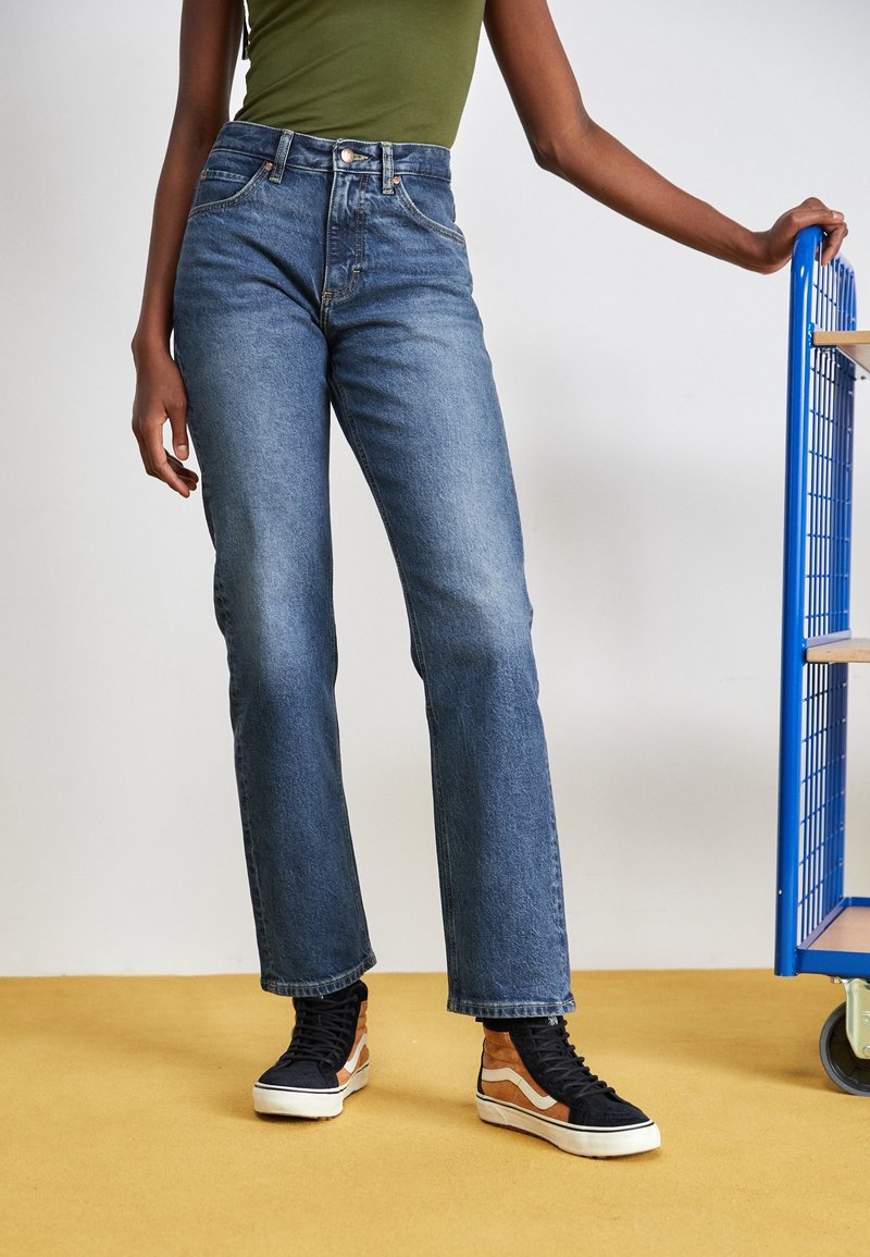 Jeans in denim blu a vita alta con design a gamba dritta, caratterizzati da cuciture a contrasto e tasche frontali, abbinati a sneaker nere e marroni.