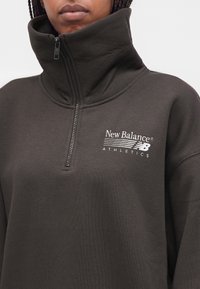 Άτομο που φοράει σκούρο γκρι πουλόβερ New Balance Athletics με φερμουάρ στο ένα τέταρτο και ψηλό γιακά, εστιάζοντας στον άνω κορμό και το λογότυπο.
