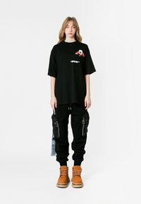 Camiseta negra de gran tamaño con diseño gráfico y bolsillo, combinada con joggers negros con cremalleras laterales y botas marrones. Textura suave.