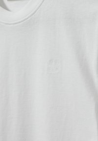 Biely bavlnený tričko s okrúhlym výstrihom a jemne reliéfne vyrazeným logom na hrudi, ktorý má hladkú textúru a štandardný strih.