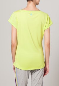 Ljusgul tränings-T-shirt med korta ärmar, lös passform och en liten blå logotyp nära halsringningen. Slät tygstruktur.