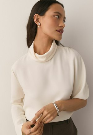 Woman with long dark hair wears a white high-neck blouse, pearl and flower earrings, a silver bracelet, and two rings while looking right.