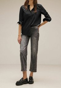 Camicia nera con bottoni, maniche arrotolate, abbinata a jeans grigi a gamba dritta e mocassini neri con zeppa, che mostrano un look casual.