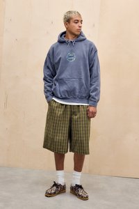 Felpa grigia con tasca frontale e testo grafico verde sul petto, abbinata a pantaloni corti a quadri verdi, calze bianche e scarpe con fantasia.