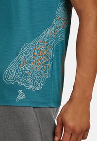 Sivuperspektiivistä katsoen henkilö, joka pitää päällään teal-väristä paitaa, jossa on valkoinen ja oranssi kontuuri-kartta -kuvio, sekä harmaat housut. Henkilön käsi roikkuu rentona sivulla.