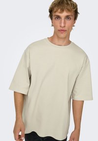 T-shirt beige à manches courtes en coton. Il présente un col rond, une coupe décontractée et une texture lisse avec un ourlet légèrement arrondi.